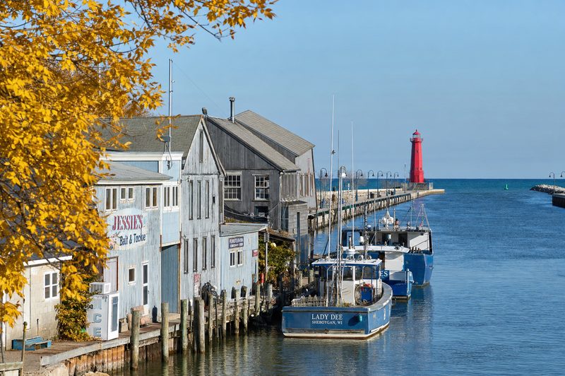 Weathered fishing shanties and boats along the Sheboygan riverfront with a red lighthouse pier beyond.